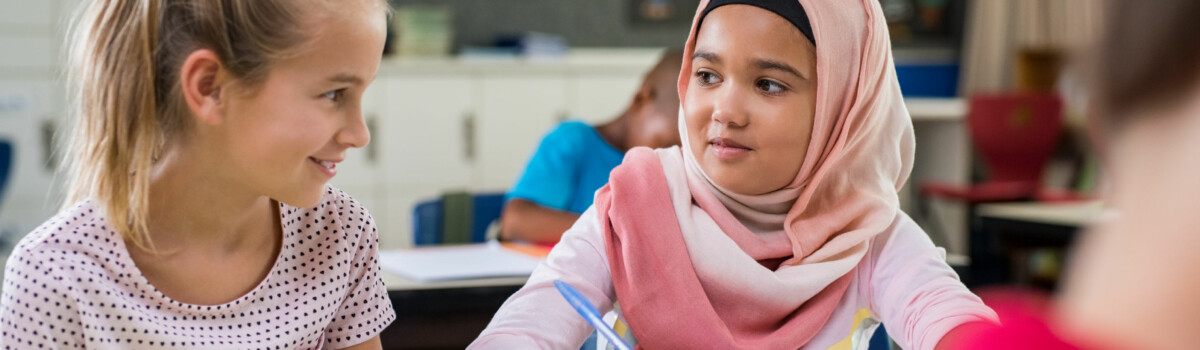 Children sat in classroom talking together while teacher completes welfare checks